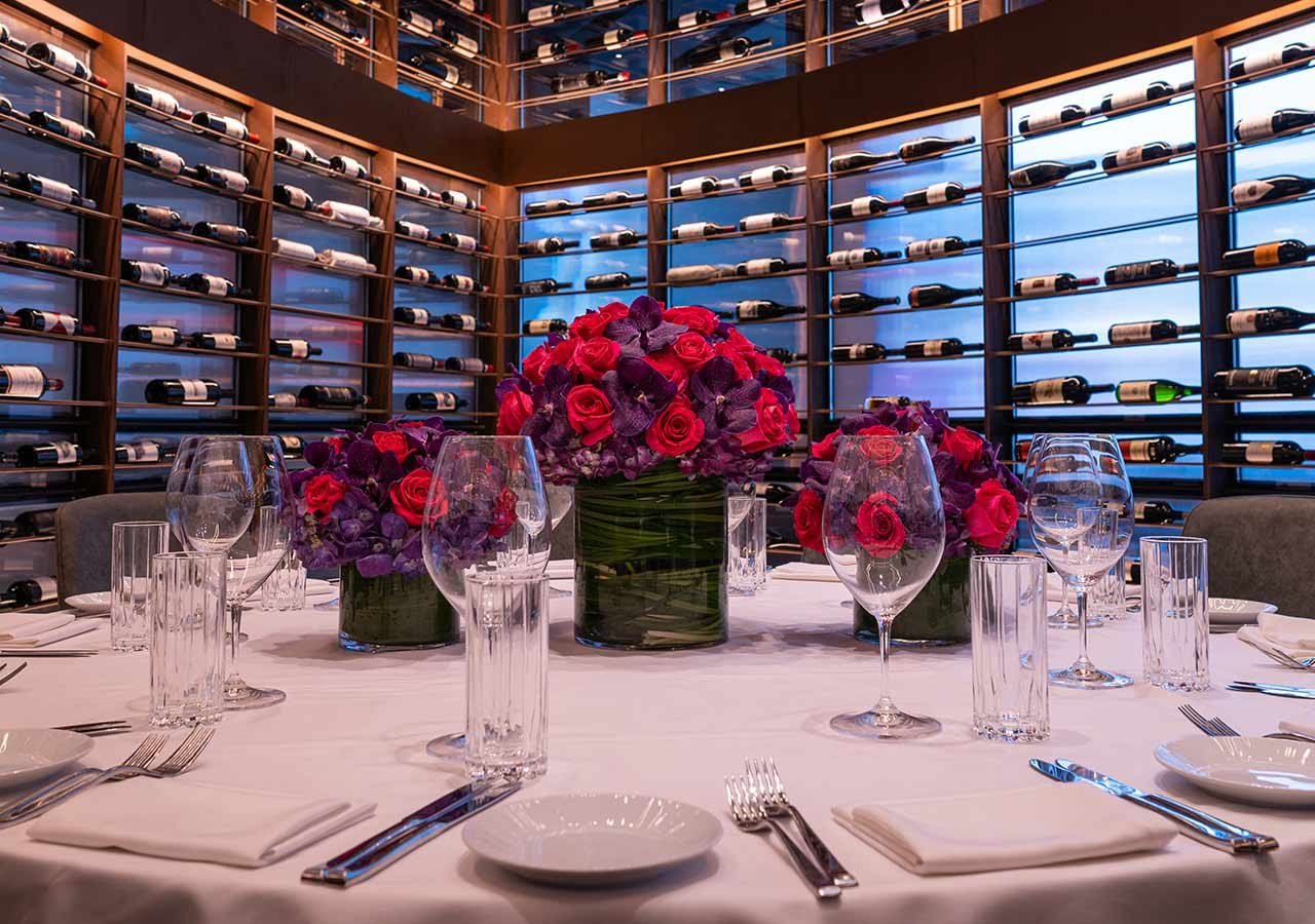 Three floral arrangements on a set table with a large assortment of wine bottles on a wine rack behind.