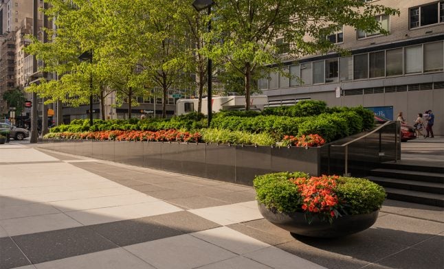 Raised commercial planter bed and a round bowl planter filled with orange impatiens and low green shrubs along a landscaped NYC sidewalk, framed by street trees and office buildings.
