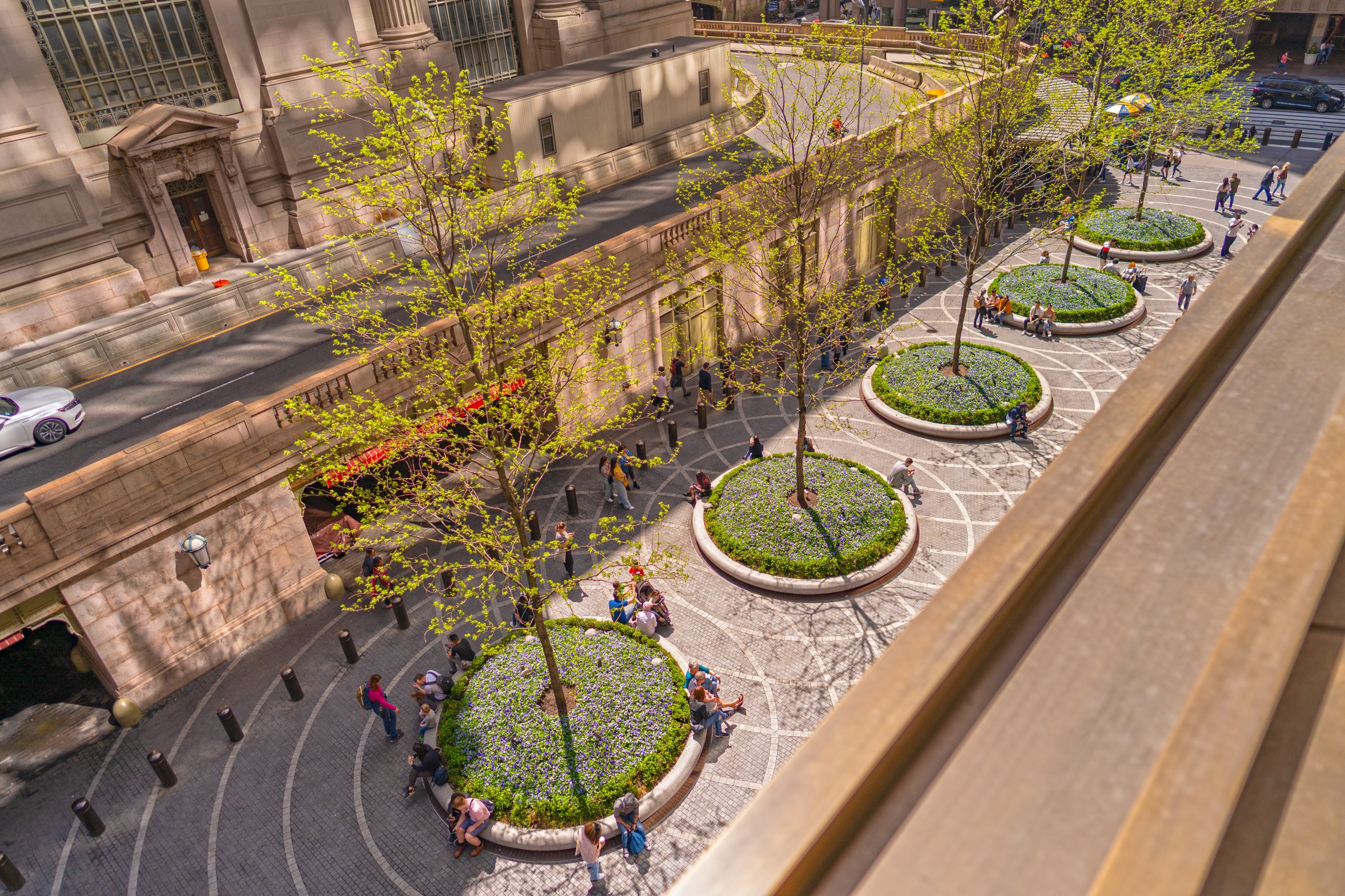 Aerial view of a city plaza with circular raised planters, leafy trees, and people sitting and walking along curved stone pathways beside a historic building.