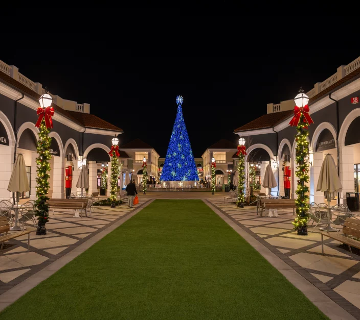 Outdoor shopping center in New York City decorated for the holidays, featuring a large illuminated blue Christmas tree at the end of a central walkway, with garland-wrapped columns topped with red bows and warm-lit storefronts on both sides