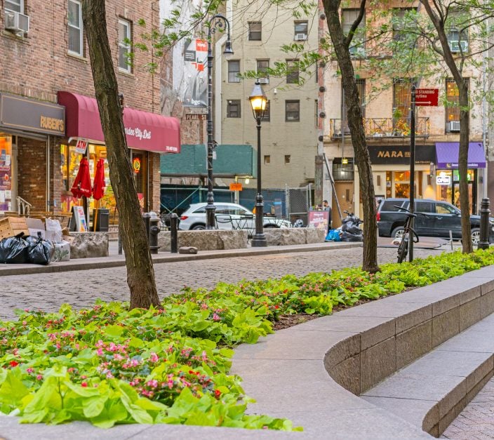 Raised planter bed with pink begonias and green foliage lining a cobblestone pedestrian plaza in an urban commercial district