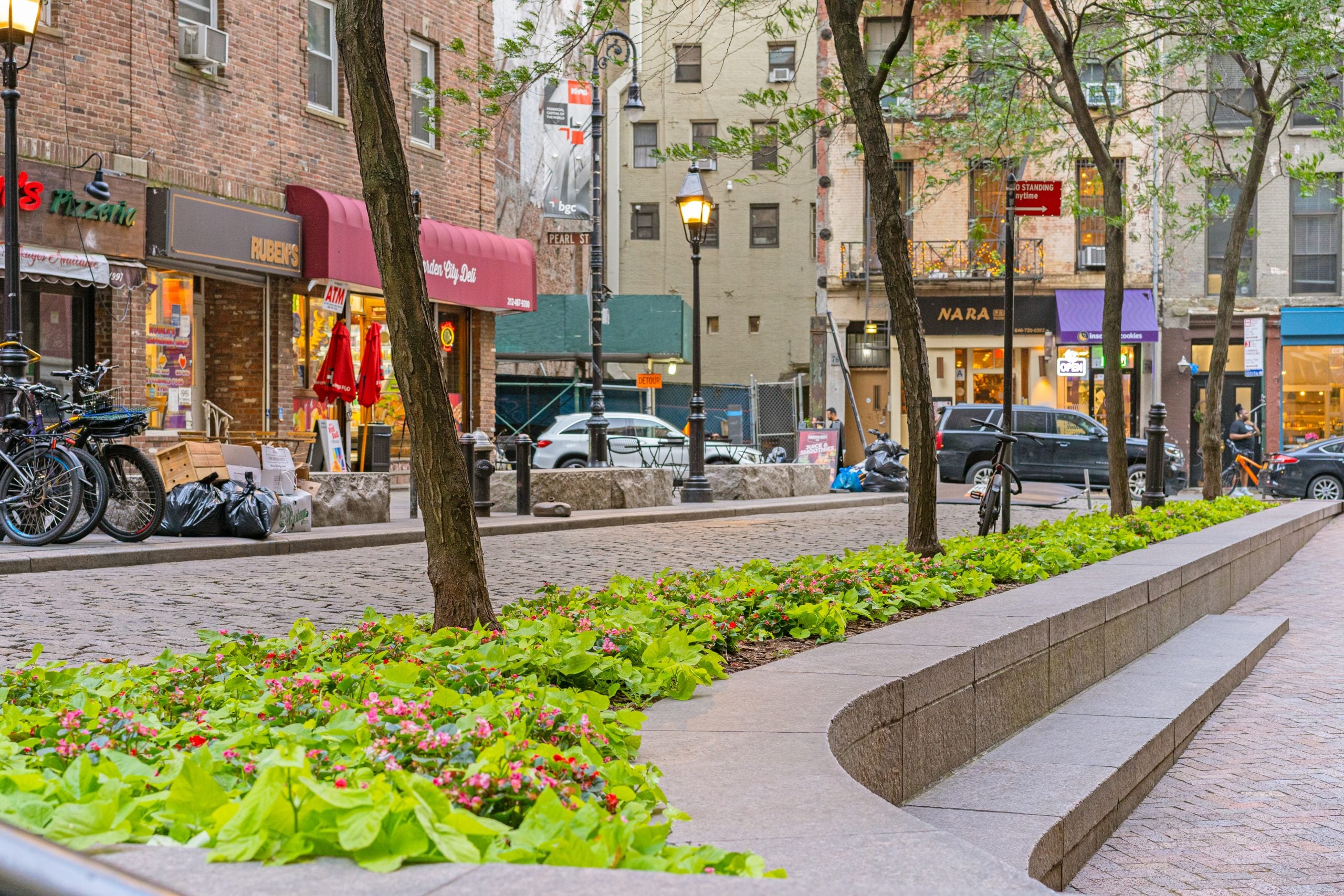 Raised planter bed with pink begonias and green foliage lining a cobblestone pedestrian plaza in an urban commercial district