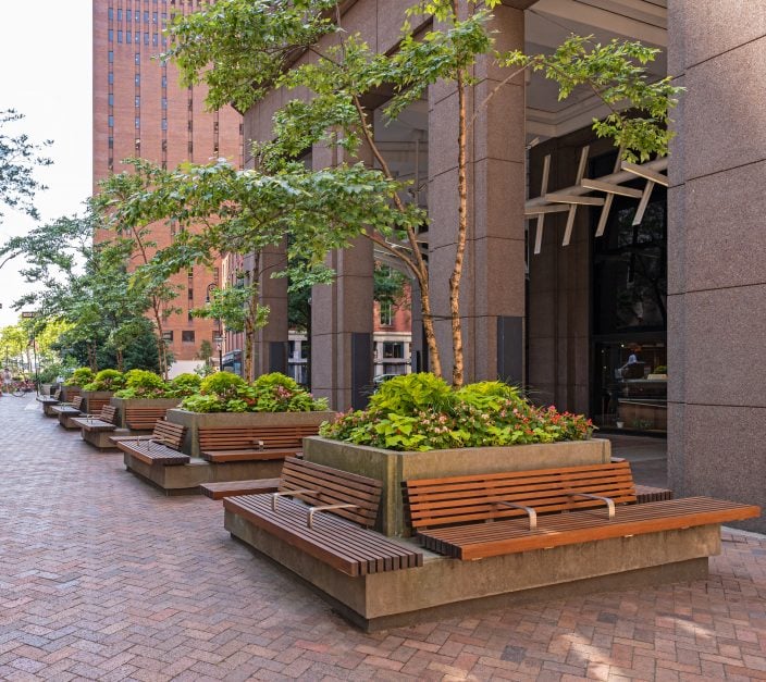 Concrete raised planters with multi-stem trees, pink begonias, and chartreuse foliage lining a brick-paved commercial plaza entrance, with integrated wood-slat benches