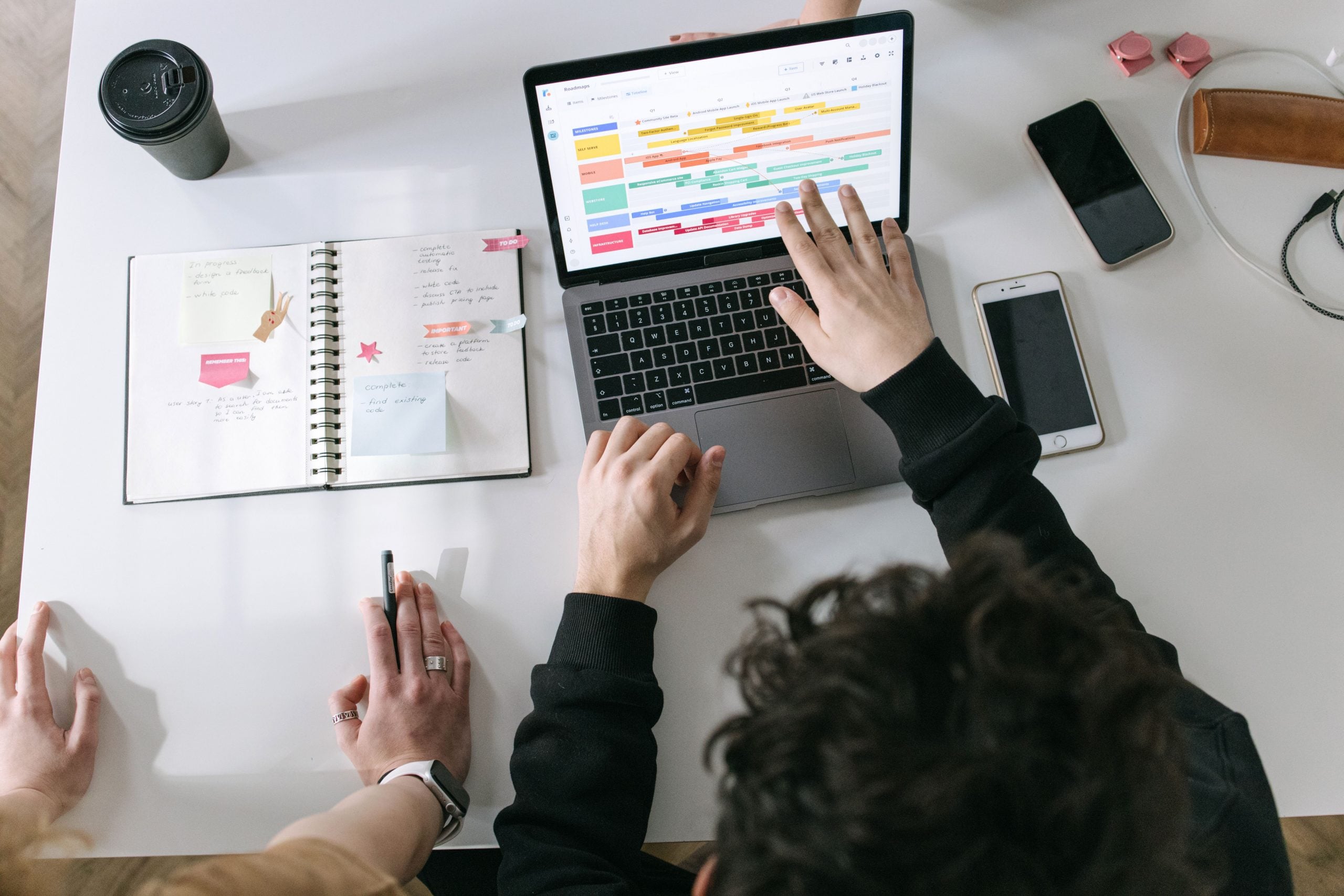 Two people planning at a desk with a laptop showing a project scheduling timeline, an open notebook, and a pen.